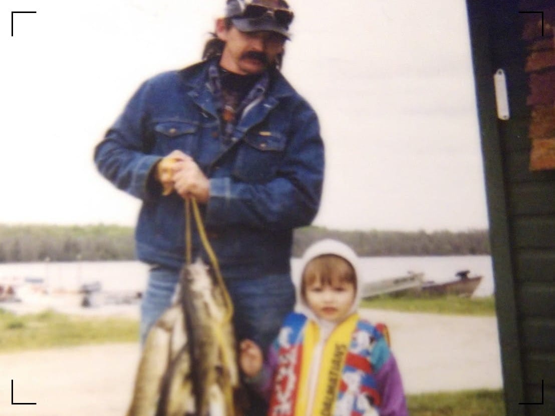 A man, 39, with long dark hair and a thick dark moustache. He holds a catch of walleye, perch, and northern pike. Next to him is a three year old in a 101 Dalmatians-themed life vest.
